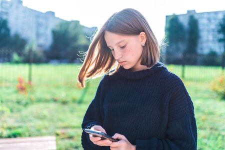 Girl teenager schoolgirl, summer city background is green grass, warm sweater, holding mobile phone, reading and writing message, online application on social networks Internet.の写真素材