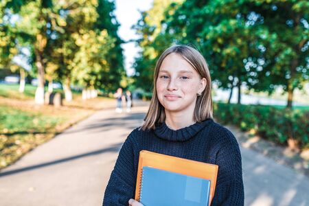 Girl teenager schoolgirl, summer city background trees and road, warm sweater, in hands of notebooks books, smiling happy, after lessons at school and institute, free space for textの写真素材