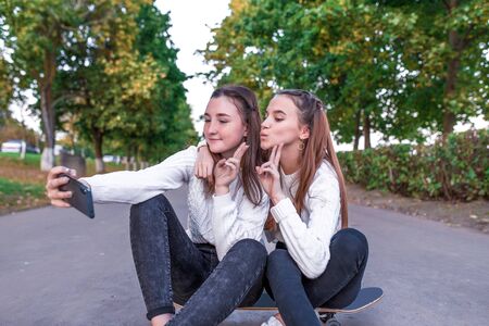 Two girls schoolgirls 12-14 years old, in summer in city park, taking selfie photo on smartphone, sitting on skateboard, casual clothes, break school and college, online official networks on Internetの写真素材