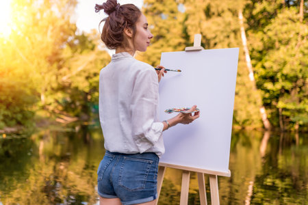 woman draws picture, view back, girl artist, summer park lake pond river white shirt denim shorts, creating creativity artistic mood. Blank white canvas, getting started. Water trees grass backgroundの写真素材