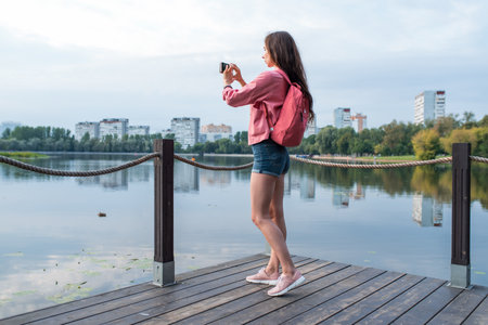girl in summer in a city park near reservoir, pond, river and lake, records video photos on smartphone. Sightseeing and travel. Landscape photography. Denim shorts jacket backpackの写真素材