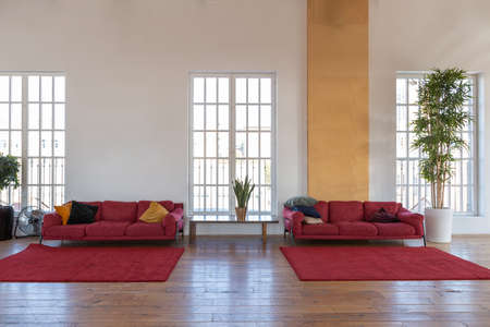 modern design of a huge white bright room with two red sofas and many large windows. full of...