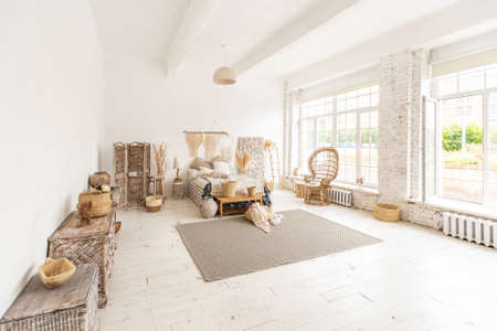 Large spacious room with a trendy loft design of sitting area. huge windows and stylish wicker light furniture inside. an abundance of ethnic decorの写真素材