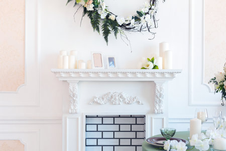 Luxury stylish bright light interior of sitting room. White walls decorated by ornament. Fireplace. Nobody inside room. Table setting by dishes, candles and flower bouquets.の写真素材