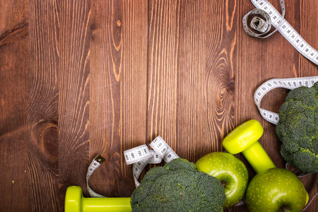 Fitness concept with dumbbells, measuring tape and fresh green vegetables on wooden backgroundの写真素材