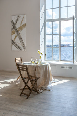 romantic table is served for two at a huge window in an open-plan apartment in a modern style with light walls and wooden floors.の写真素材