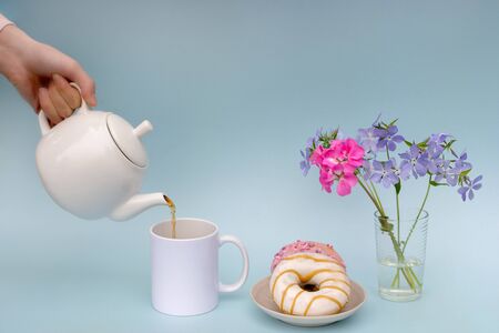 A womans hand pours tea from a white teapot into a white mug, a plate with 2 doughnuts, and a vase of blue flowers. The photo is made in pastel colors on a blue background. The concept of Breakfastの写真素材