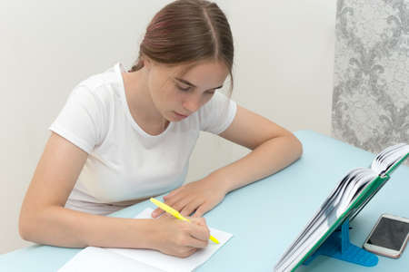 A beautiful girl teenager sits at the table and writes in a clean notebook. Concept - homework,...