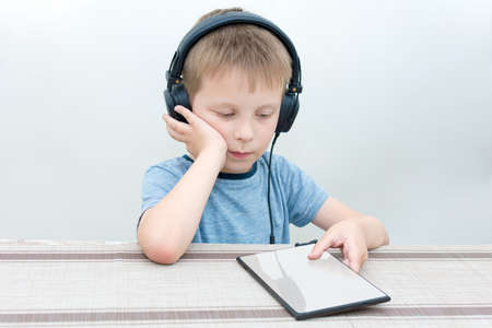 a cute Boy in headphones sits at a table and looks with interest at something on the tablet. The idea is to listen to music or an audiobook, to learn something new and interesting.の写真素材