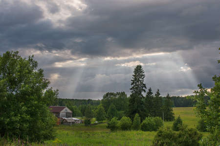 The sun's rays break through thick dark clouds over the village, field and forest. Dawn, sunset. Horizontal photoの写真素材