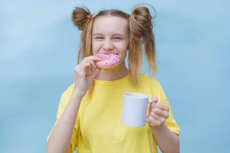 funny, cheerful girl bites off a donut and holds a white mug with a drink in her hands. Girl in a yellow T-shirt on a blue background.Horizontal photoの写真素材