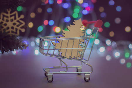 A close-up of a mini cart with a wooden Christmas tree in the background of Christmas lights. Concept - holiday shopping, delivery, Christmas sales, preparing for Christmas, approaching the new yearの写真素材