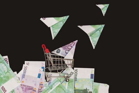 against a black background, a mini-cart from a supermarket stands on money, and planes from euro...