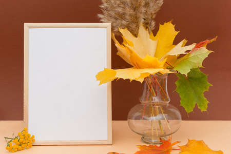 Wooden photo frame mockup stands upright on a brown background. Nearby is a vase with autumn leaves, fluffy branches and flowers. Photo layout, copy spaceの写真素材