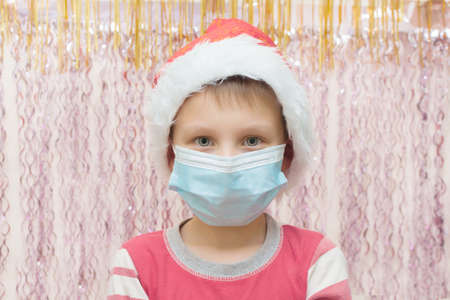 A boy celebrates Christmas during the covid-19 pandemic in a protective medical mask. New Year holidays holiday. festive mood in 2021. Close-up, against the background of New Year's tinselの写真素材