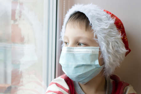 A close-up portrait of a preschool boy wearing a medical mask and a New Year's cap. A boy looks out...