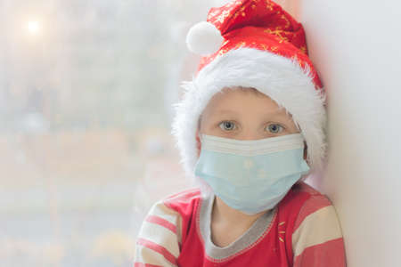 preschooler in a medical mask and a New Year's cap looks at the camera against the background of a window. Waiting for Christmas and New Year in modern reality and the covid-19 pandemic. Soft focusの写真素材