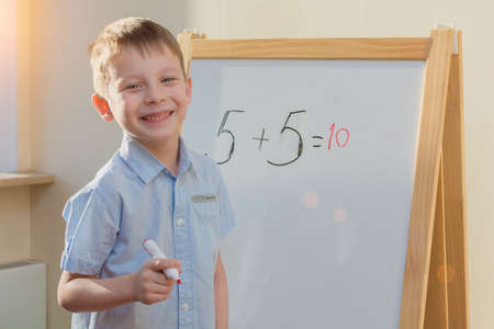 happy, smiling preschooler stands with a red marker in his hands at a white school board with a...