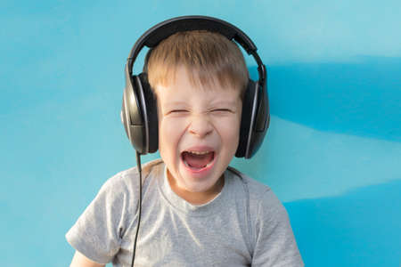child in headphones, on a blue background, portrait. A boy with an open mouth and a funny facial expression. Horizontal photoの写真素材
