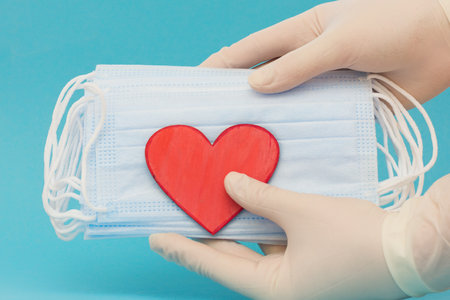 Hands in medical gloves hold a pack of medical masks and a small red heart on a blue background....