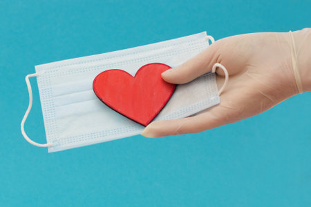 a hand in a medical glove extends a mask with a red heart. Concept - concern for the safety of others, Valentine's Day, horizontal photo, close-upの写真素材