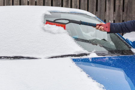 A womans hand with a special brush sweeps snow from the car. Removes the effects of a blizzard. A hand sweeps snow from the windshield of a car. Horizontal photo.の写真素材
