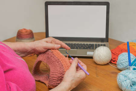 Close-up - the hands of a woman who is crocheting something, next to a laptop. Online training, online training, online needlework lessons. Horizontal photoの写真素材