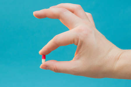 a beautiful red and white pill clamped in his fingers. Shows a pill. A pill for all diseases, vitamin, food supplement, Horizontal photo, close-up. Blue background.の写真素材