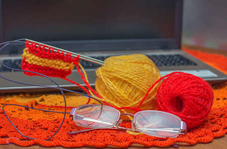 on a red knitted napkin - balls of thread and glasses. In the background is a laptop. Concept -...