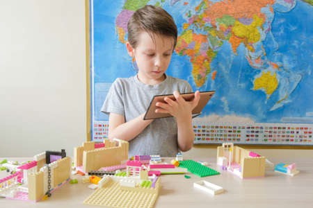 boy with a tablet in his hands on the background of the world map sits at the table, the child looks...