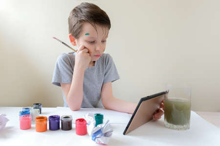 A preschool boy sits at a table with a brush in his hands. The child looks at the tablet. Learns to...