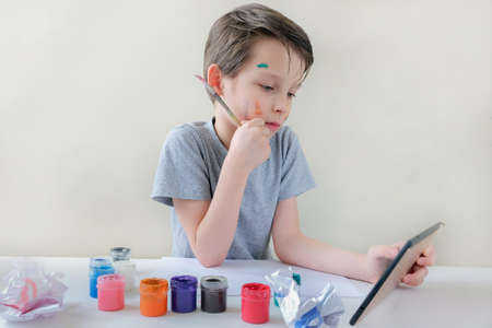 boy with a tablet in his hands sits at the table, paints and paper on the table, learning to draw...