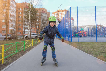 A preschool boy learns to roller-skate on an asphalt path. Child in a protective helmet and special...