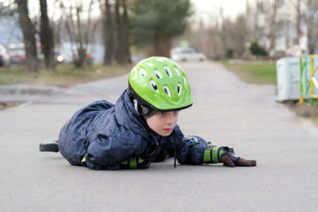The boy lies on the asphalt in a green helmet and special hand protection. The child fell while...