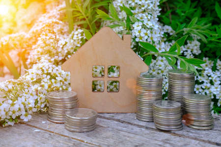 a small wooden model of a house among blooming white flowers of greenery and stacks of coins. Concept - buying real estate, mortgage, renting a house, investing in real estate. Housing mortgage rate.の写真素材