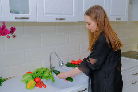 A beautiful teenage girl in pajamas washes vegetables under the tap in the kitchen. Good morning, In the hands of a cucumber. Healthy breakfast, healthy lifestyle. Proper nutrition. Horizontal photoの写真素材