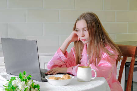 A beautiful sad girl is sitting with a laptop in the kitchen. A serious student doing her homework, working on a research project on the Internet, doing her homework. Online education, home teaching.の写真素材
