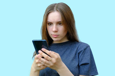 young beautiful girl with brown eyes isolated on a blue background. The teenager looks at the phone with a surprised expression on his face. Horizontal photo. Concept - bad news on the phoneの写真素材