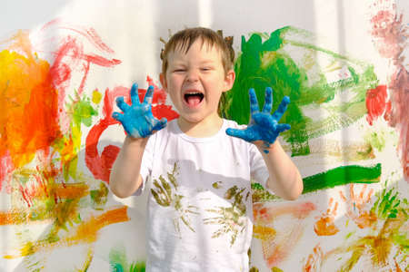 a boy in a white T-shirt, stained with paint, and dirty blue hands, stands against the background of a multi-colored wall. The child painted with his hands. A funny, lovable boy, looks into the frame.の写真素材