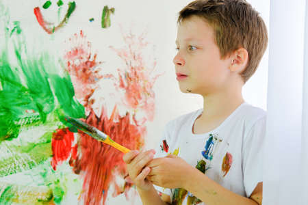 a boy in a dirty T-shirt stands against the background of a multi-colored wall with a brush in his hands. The preschooler painted with paints on the wall. the child is upset, offended, crying.の写真素材