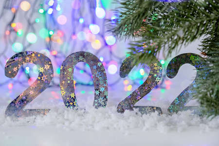 The year 2022 stands against the background of Christmas lights and fir trees. The numbers are brilliant, with a pattern of stars. Snow lies in front of the numbers. New Years, festive mood.の写真素材