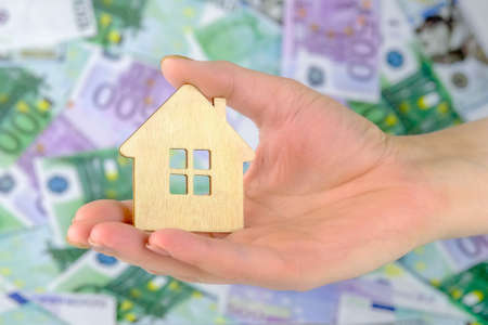 in a female hand a small wooden house on the background of paper Euro bills. Concept - buying real estate, selling, investing in real estate, home insurance.の写真素材