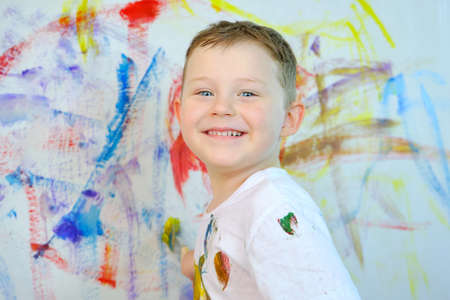 A handsome blue-eyed boy is looking at the camera and smiling. The child draws with paints on the wall. Childrens creativity. Horizontal photo.の写真素材