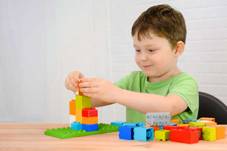 A boy in a green T-shirt sits at a table in the room and collects a constructor set from colorful...
