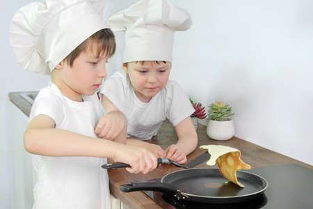 two boys 4 and 7 years old cook pancakes in a pan in the kitchen. The brothers learn to cook on their own. Children in the form of cooks, caps and apron. serious expression.の写真素材