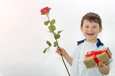 A happy smiling boy 4-5 years old holds a gift and a red rose in his hands. Child on the background of a white brick wall. Surprise idea for mom for mothers day, birthday.の写真素材