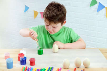 a boy of 4-5 years old sits at a table in a room and paints wooden Easter eggs with green paint. A...