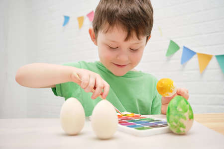 cute smiling boy 4-5 years old paints an easter egg with yellow paint. The child is busy with...