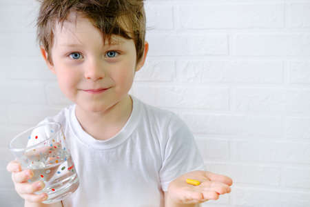 the boy is looking at the camera, holding a glass of water and a yellow pill in his hand. The child is going to take medication. Horizontal photo, portrait of a child.の写真素材