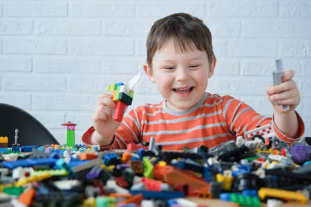 a happy child sits at a table in front of scattered toy building blocks. The boy holds the details of the designer in his hands, laughs, has fun.の写真素材